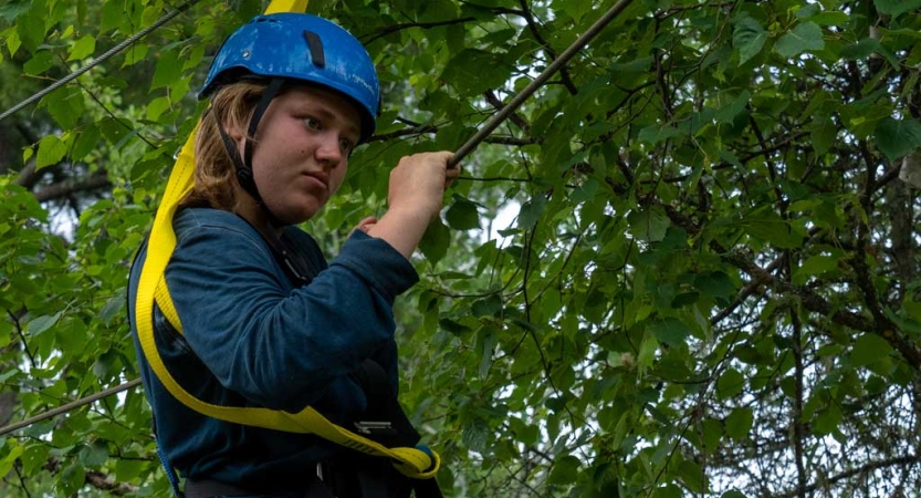 A person wearing safety gear is secured by ropes as they balance on an obstacle on a high ropes course. 